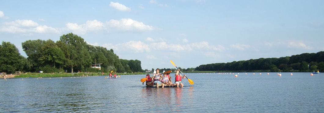 Fünf Jungen paddeln auf einem selbstgebauten Floss auf einem See. Die Jungen tragen Schwimmwesten und haben Paddel in den Händen.
